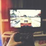 Mão segurando um controle de videogame diante de uma televisão, em ambiente interno.