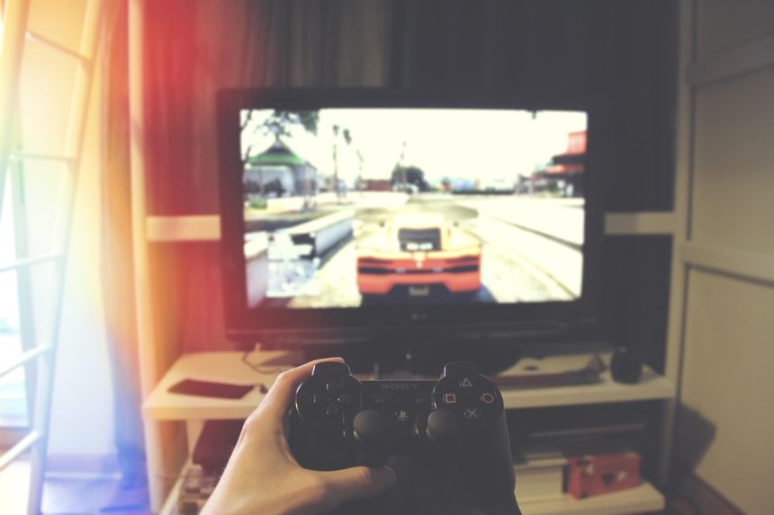 Mão segurando um controle de videogame diante de uma televisão, em ambiente interno.